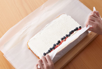ホイップクリームを作る。ボウルに生クリーム、グラニュー糖を入れ、泡立て器で8分立てにする（泡立て器ですくって角が残るくらい）。いちごは縦半分に切る。まな板などにオーブン用シートを敷いて生地をのせ、ホイップクリームを均一に塗る。手前から1/3のところにいちご、ブルーベリーを並べる。