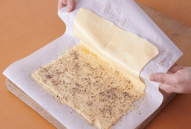 横向きにして全体に溶き卵を塗り、半分に粉チーズと粗挽き黒こしょうをふる。オーブン用シートごとパイシートを持ち上げ、半分に折りたたむ。